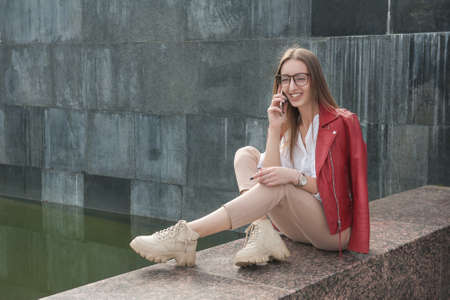 Beautiful young fashion stylish woman talking on the phone and laughing sit on parapet near wall city backgroundの写真素材