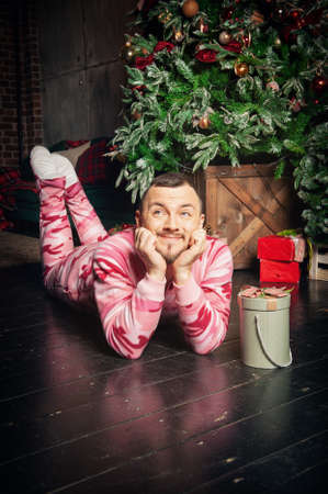 Cheerful funny man in pink sleepwear lying down on the floor near decorated fir tree and Christmas presentの写真素材