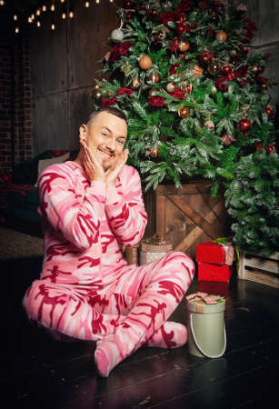 Cheerful surprised funny man in pink sleepwear sitting on the floor near decorated fir tree and Christmas presentの写真素材
