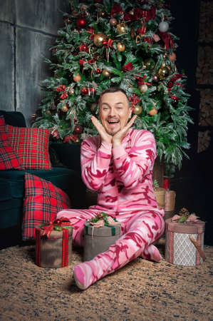 Cheerful surprised funny man in pink sleepwear sitting on the floor near decorated fir tree and Christmas presentの写真素材