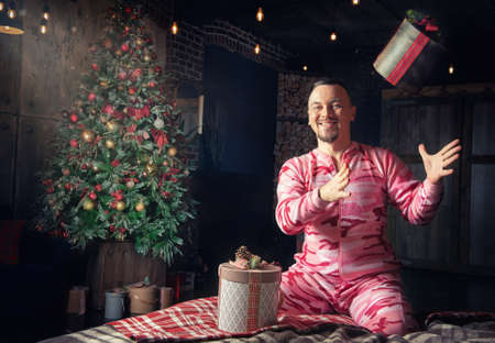 Cheerful funny man in pink sleepwear sitting on the bed near decorated fir tree and throws up...