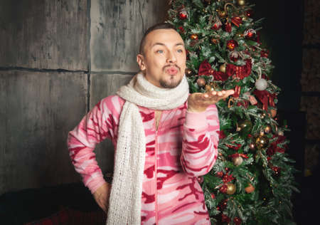 Cheerful funny happy man in pink sleepwear stand near decorated fir tree and sending kissの写真素材