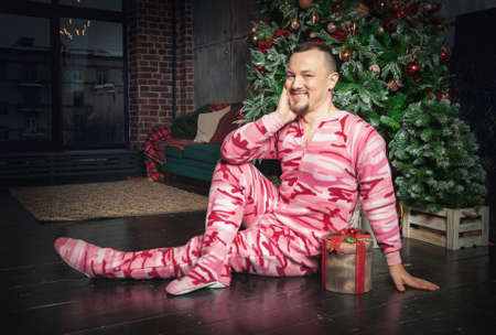 Cheerful happy funny man in pink sleepwear sitting on the floor near decorated fir tree with Christmas presentの写真素材