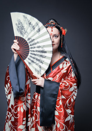 Beautiful young woman in red traditional Chinese costume with hand fan on dark backgroundの写真素材