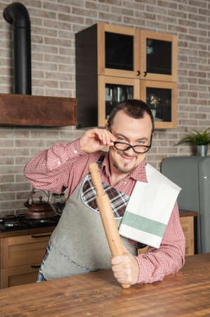 Funny handsome young smiling man wearing pinafore with towel and eyeglasses on the kitchenの写真素材