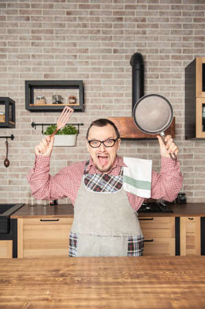 Funny handsome screaming angry man wearing pinafore with kitchen utensilsの写真素材