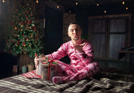 Cheerful funny man in pink sleepwear sit on the bed near decorated fir tree, send kiss and hold Christmas presentの写真素材