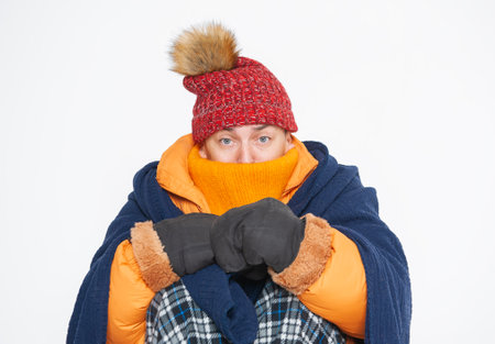 Frightened handsome man wearing in red hat, yellow down jacket and plaid feeling cold sitting on white background. gas crisis conceptの写真素材
