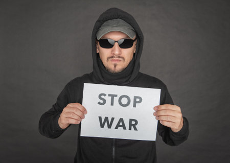 Man in the black hoody with hood wearing sunglasses holding stop war on white sheet of paper on dark backgroundの写真素材