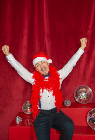 Happy smiling man wearing Santa hat sitting on the red background with hands up. new years styleの写真素材