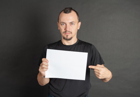 Young handsome man holding empty white sheet of paper and showing on it by his finger on dark backgroundの写真素材