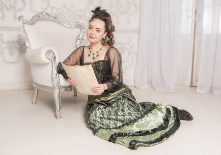 Beautiful young woman wearing green medieval vintage Victorian Style dress read letter on the floor near armchairの写真素材