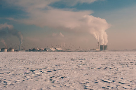 Winter landscape with snow covered frozen ice, flying birds, city and pipes with thick steamの写真素材