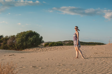 Beautiful woman wearing dress and sunglasses walking on the sand against sky backgroundの写真素材