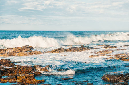 Sea waves crashing on rocks in sunny day against blue cloudy skyの写真素材