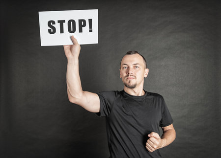 Young serious man holding white sheet of paper stop signunder his head on dark background