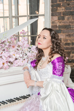 Young beautiful woman in fantasy white and purple rococo style medieval dress sitting near piano with pink flowersの写真素材