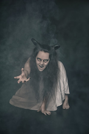 Terrible screaming ghost with horns reach put hand on dark background with smokeの写真素材