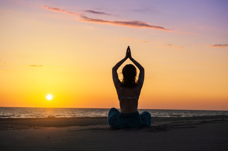Beautiful young woman exercising yoga while doing meditation at beach during sunrise or sunset sitting on the sand against sea water and skyの写真素材