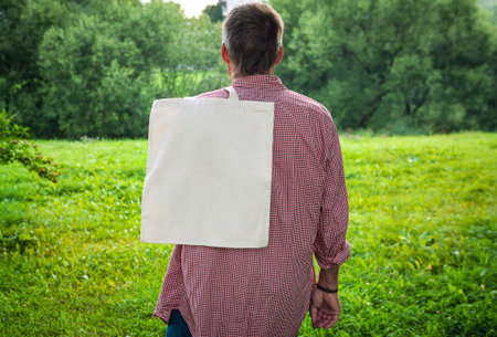 Man wearing checkered shirt and jeans holds an eco-friendly shopping handbag, standing outdoors...