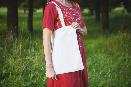 Woman in a long red dress holds an eco-friendly shopping handbag, standing outdoors among grass and...