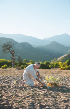 Man kneels on the desert sand tending to a small plant. Quiet wilderness surrounded by mountains offers a peaceful atmosphereの写真素材