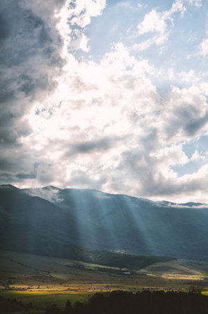 Sun rays breaking through clouds, illuminating a lush valley and mountainsの写真素材