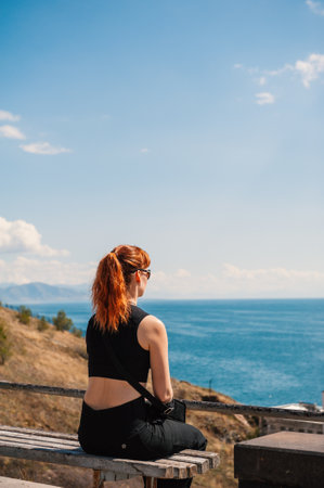 Red-haired woman sitting on a bench, enjoying a serene seaside view on a sunny dayの写真素材