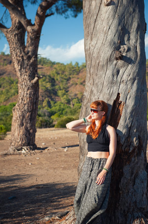 Stylish redhead beautiful woman with sunglasses leans against a rustic tree in vibrant sunlightの写真素材