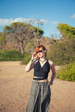Beautiful redhead woman standing confidently in a sunny landscape, bohemian outfitの写真素材