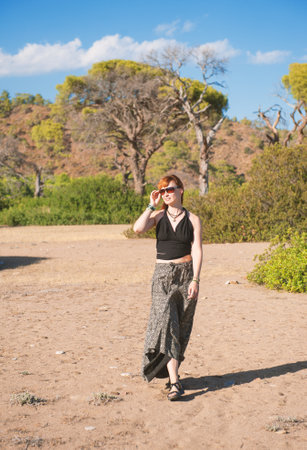 Beautiful redhead woman walking confidently through a sunny forest landscape, bohemian outfitの写真素材