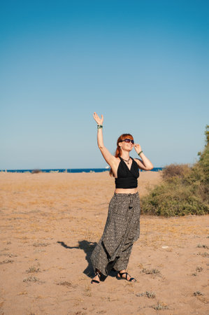 Stylish beautiful redhaired woman in sunglasses enjoys a tranquil moment by the ocean and waves her handの写真素材