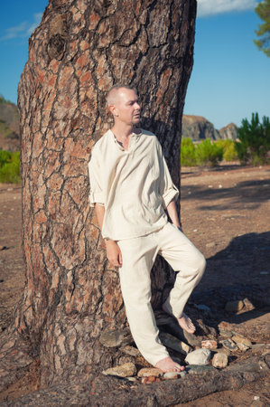 Mature man leans against a large tree in a calm forest setting. Sunlight casts soft shadows on his peaceful figureの写真素材