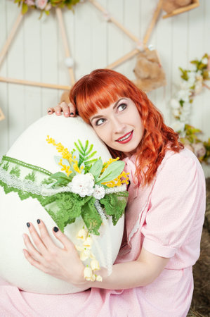 Cheerful redhead beautiful woman hugging a beautifully decorated Easter egg with floral lace detailsの写真素材