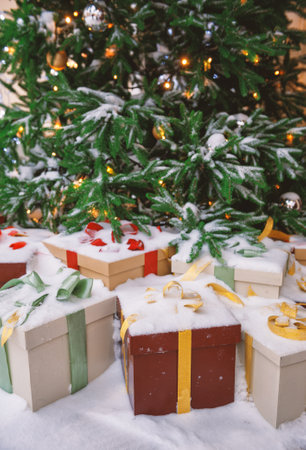 Snow-covered Christmas tree with glittering ornaments, festive lights and gift boxes wrapped in ribbons under itの写真素材