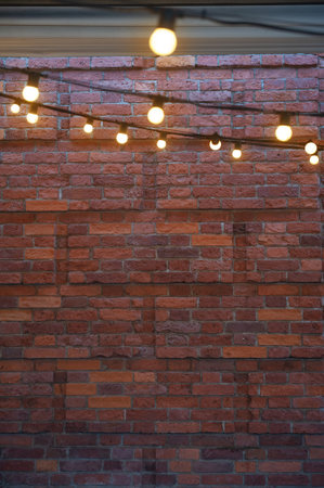 Cozy glowing string lights on a textured red-brick wall backdrop. Warm illumination creates a trendy urban ambiance on rustic bricksの写真素材