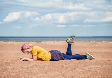 Cheerful young woman with pink hair having fun and lying down on the sand outdoorの写真素材