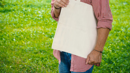 Man wearing checkered shirt and jeans holds an eco-friendly shopping handbag, standing outdoors among grass and trees. The image features empty copy space for mockup and business useの写真素材