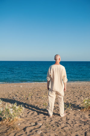 Barefoot mature man in white linen gazes at the serene ocean horizonの写真素材