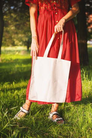 Woman in a long red dress holds an eco-friendly shopping handbag, standing outdoors among grass and trees. The image features empty copy space for mockup and business useの写真素材