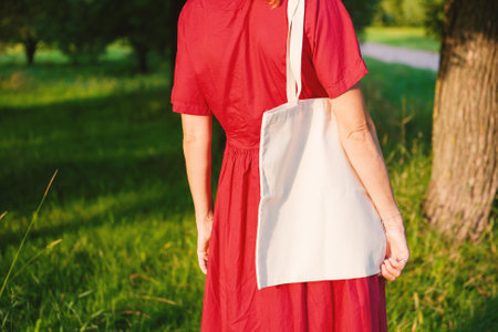 Woman in a long red dress holds an eco-friendly shopping handbag, standing outdoors among grass and trees. The image features empty copy space for mockup and business useの写真素材