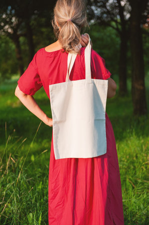 Woman in a long red dress holds an eco-friendly shopping handbag, standing outdoors among grass and trees. The image features empty copy space for mockup and business useの写真素材