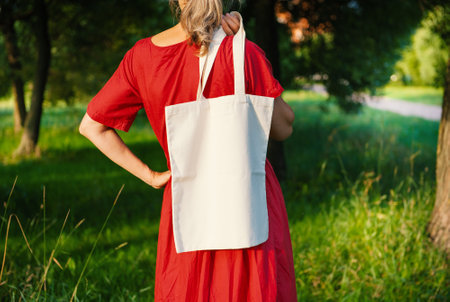 Woman in a long red dress holds an eco-friendly shopping handbag, standing outdoors among grass and trees. The image features empty copy space for mockup and business useの写真素材