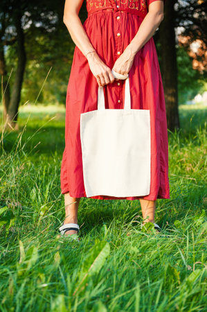 Woman in a long red dress holds an eco-friendly shopping handbag, standing outdoors among grass and trees. The image features empty copy space for mockup and business useの写真素材