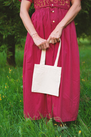 Woman in a long red dress holds an eco-friendly shopping handbag, standing outdoors among grass and trees. The image features empty copy space for mockup and business useの写真素材