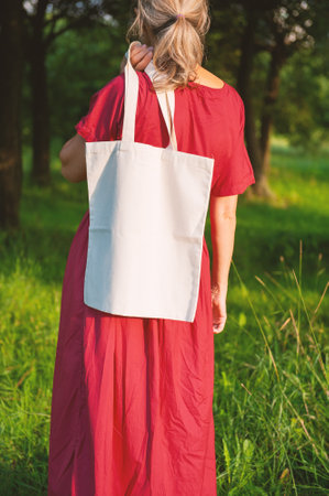 Woman in a long red dress holds an eco-friendly shopping handbag, standing outdoors among grass and trees. The image features empty copy space for mockup and business useの写真素材