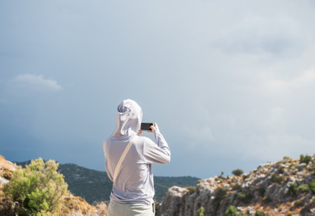 Traveler captures mountain scenery under moody cloudy sky. Solitary figure with hood photographs...