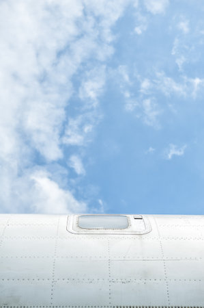 Blue sky and clouds frame the aircraft exterior of airplane fuselage with visible window and metallic texture.の写真素材