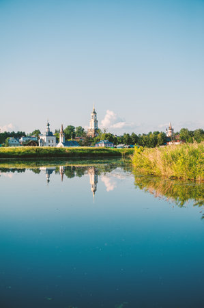 Orthodox church with green houses reflected in calm river water. Peaceful countryside view with...