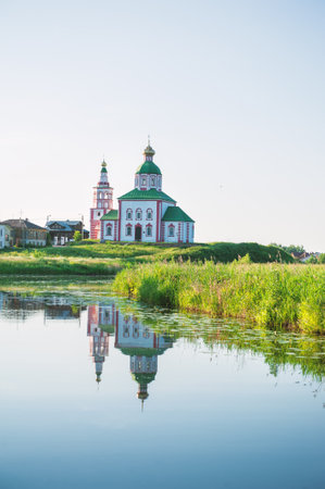 Orthodox church with green houses reflected in calm river water. Peaceful countryside view with religious architecture and lush grassの写真素材